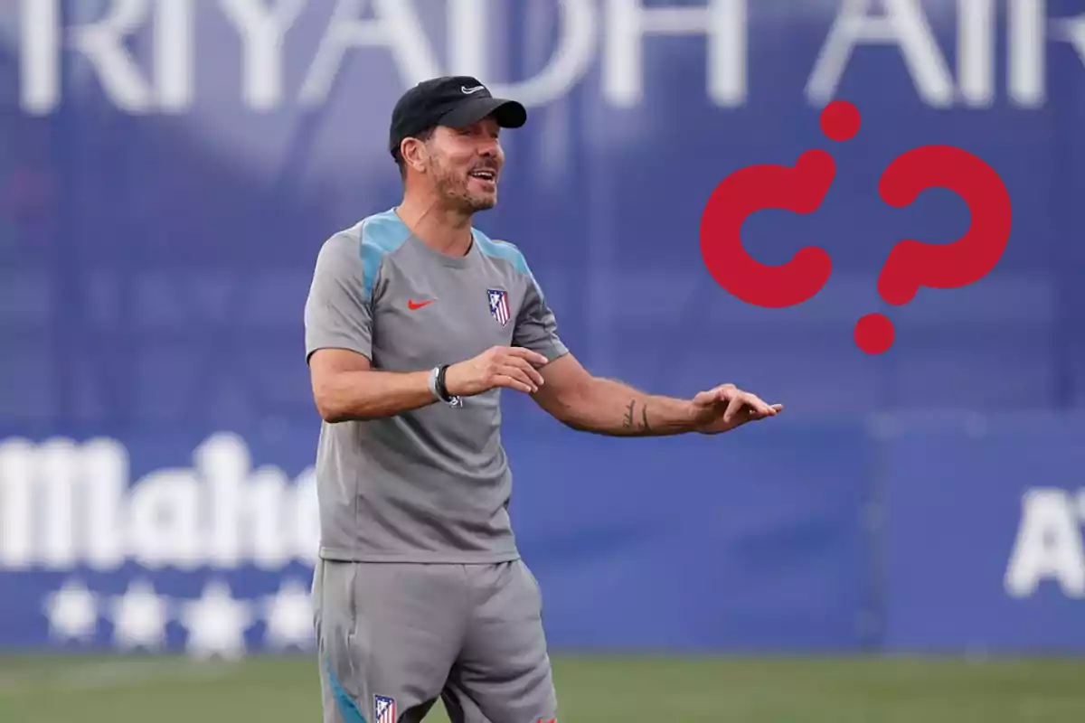 Un entrenador de fútbol con uniforme gris y gorra negra gesticula durante un entrenamiento en un campo de fútbol.