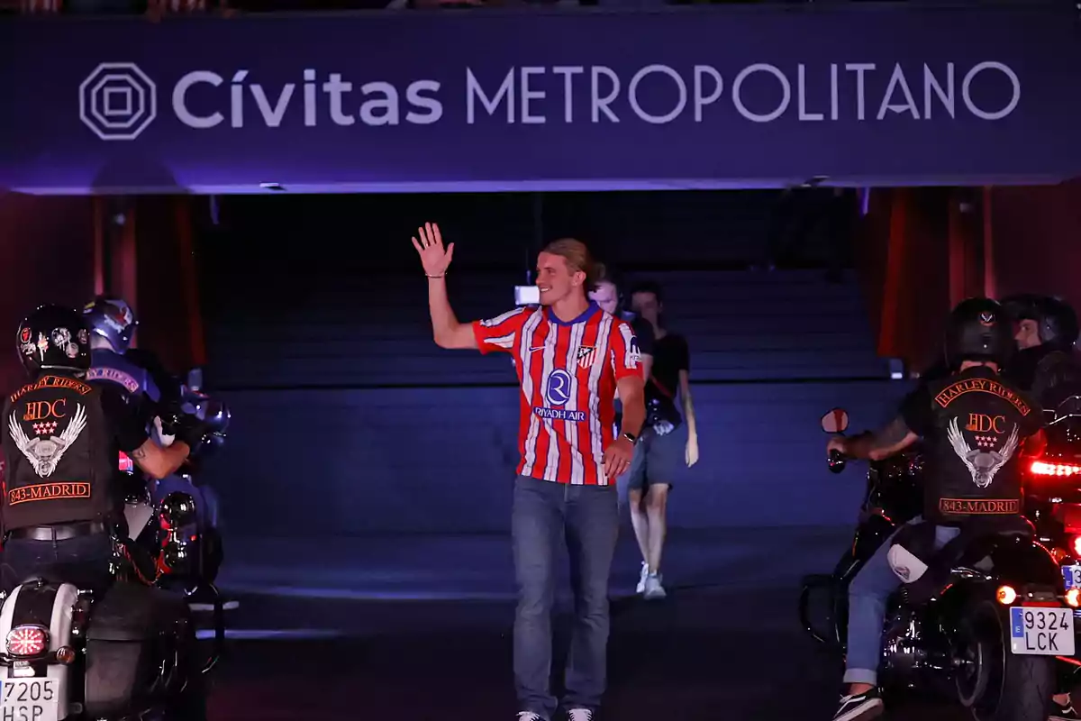 Un hombre con una camiseta de rayas rojas y blancas saluda mientras camina por un túnel en el estadio Cívitas Metropolitano, rodeado de motociclistas.