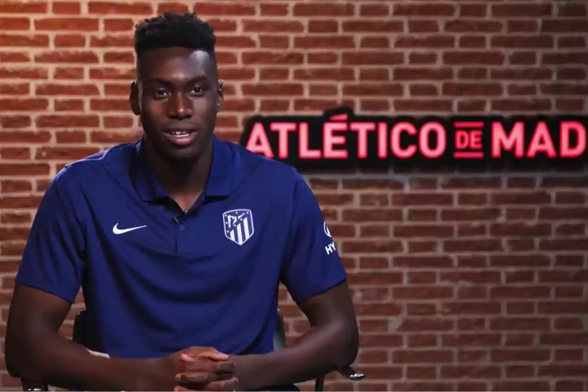 Un jugador del Atlético de Madrid con una camiseta azul está sentado frente a una pared de ladrillos con el logo del equipo en el fondo.