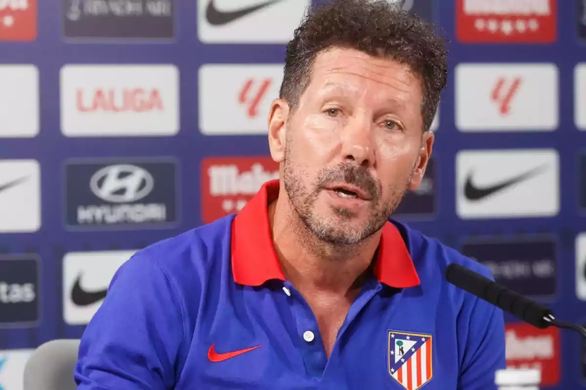 Hombre con barba en una conferencia de prensa con fondo de logotipos deportivos.