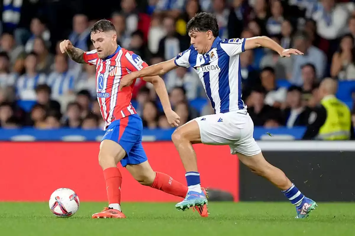 Dos jugadores de fútbol compiten por el balón en un partido, uno con uniforme rojo y blanco y el otro con uniforme azul y blanco.