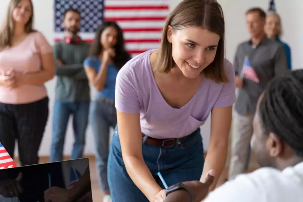 Smiling young woman handing a document to a seated person while others wait in line with a United States flag in the background Smiling young woman handing a document to a seated person while others wait in line with a United States flag in the background