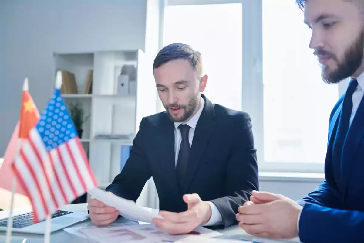 Two businessmen in suits review documents in an office with United States and China flags on the table. Two businessmen in suits review documents in an office with United States and China flags on the table.