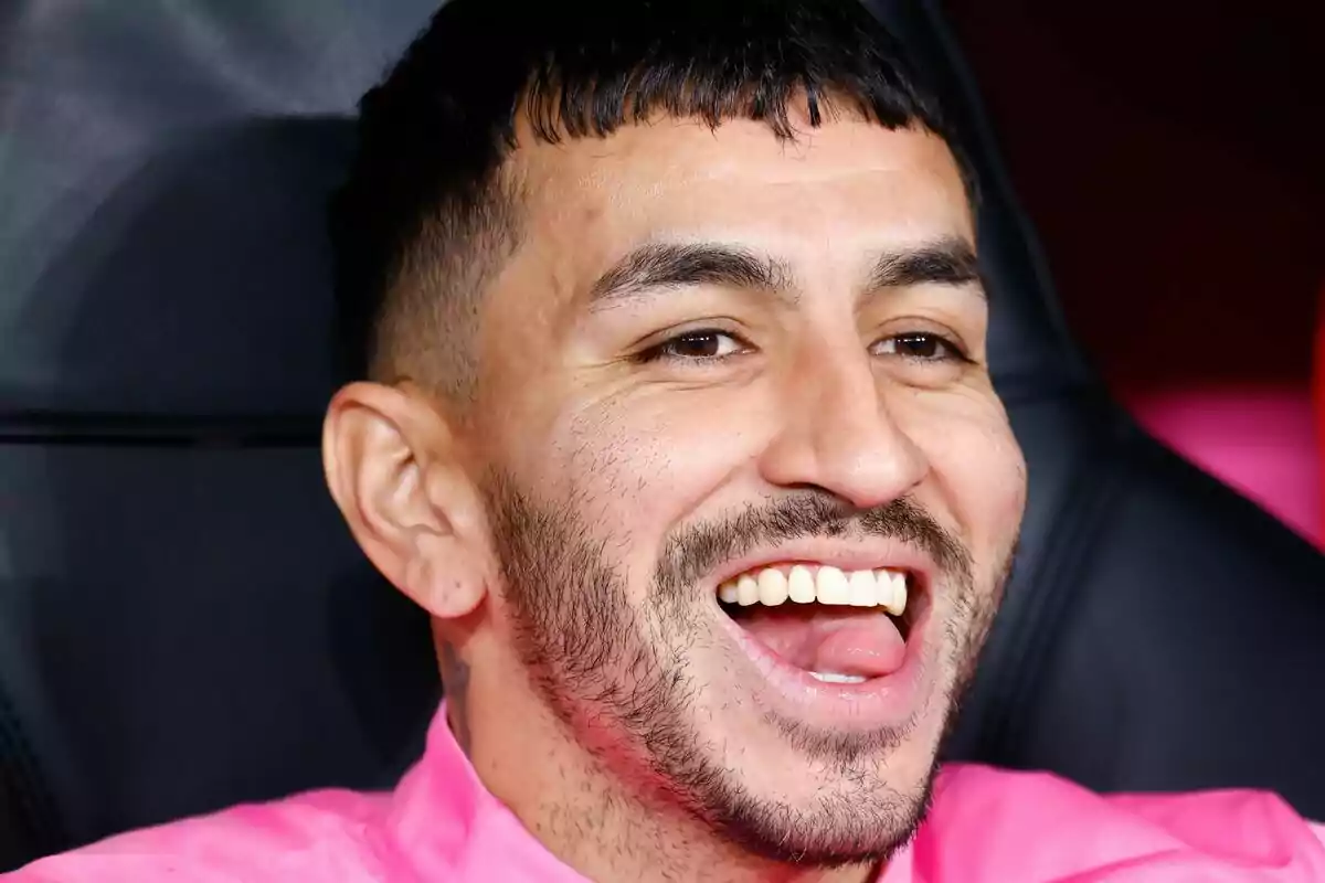 Hombre joven con barba corta y cabello oscuro sonríe ampliamente mientras viste una camiseta rosa