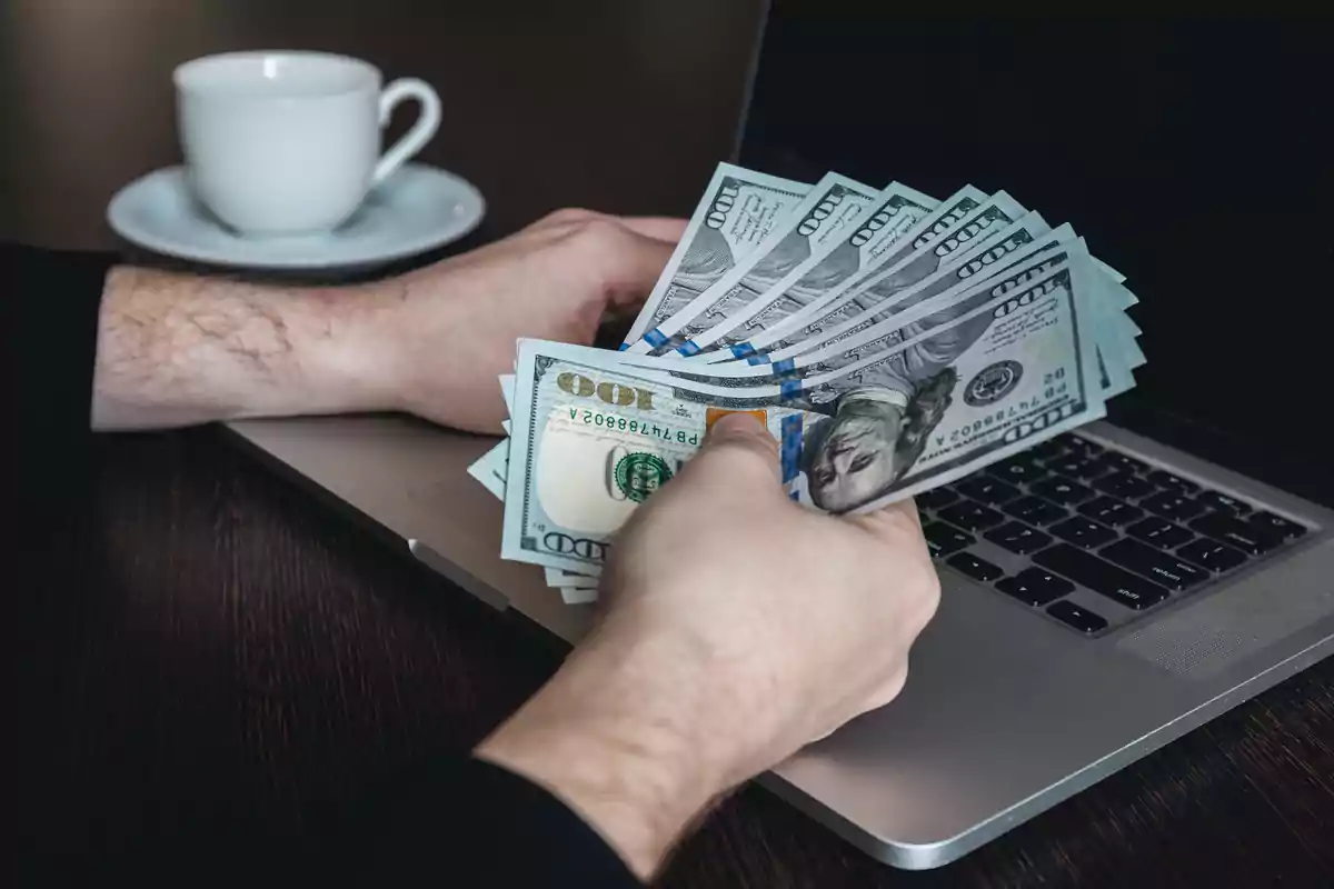 Persona sosteniendo varios billetes de cien dólares frente a una computadora portátil con una taza de café al fondo Persona sosteniendo varios billetes de cien dólares frente a una computadora portátil con una taza de café al fondo