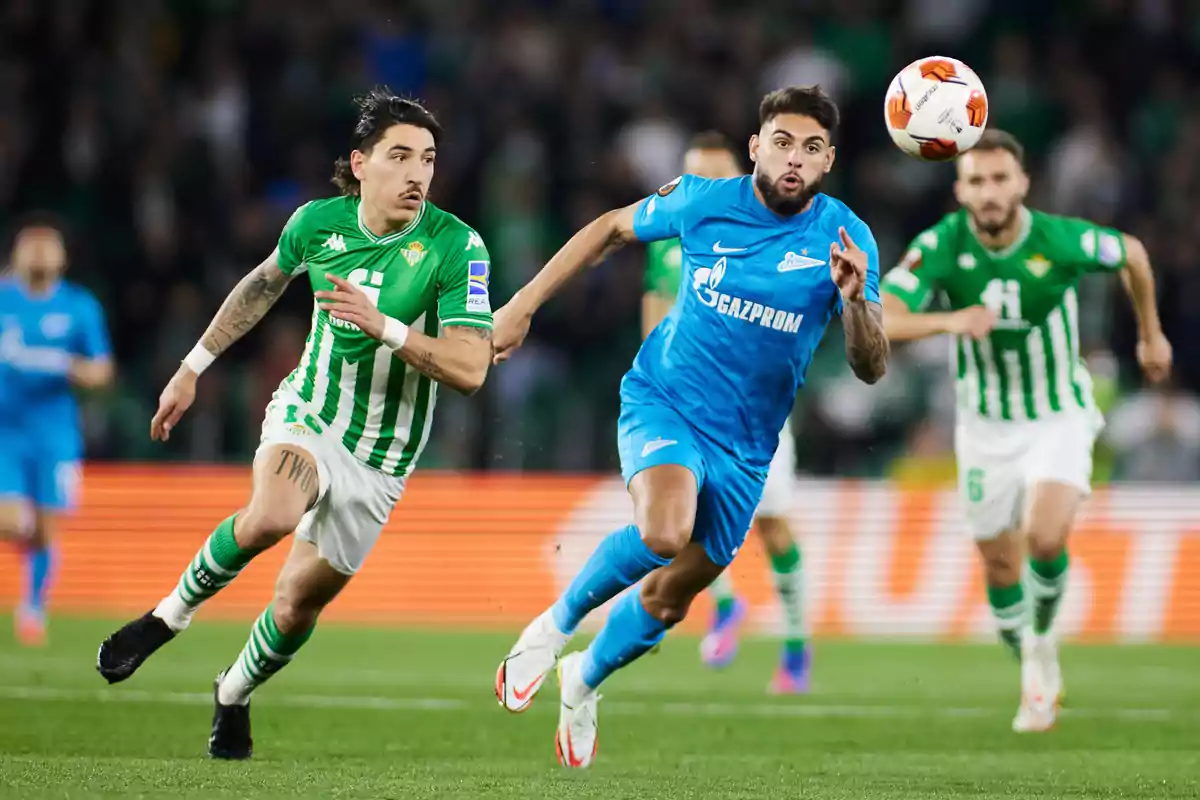 Dos futbolistas corren tras el balón durante un partido entre un equipo de uniforme verde y blanco y otro de uniforme azul Dos futbolistas corren tras el balón durante un partido entre un equipo de uniforme verde y blanco y otro de uniforme azul