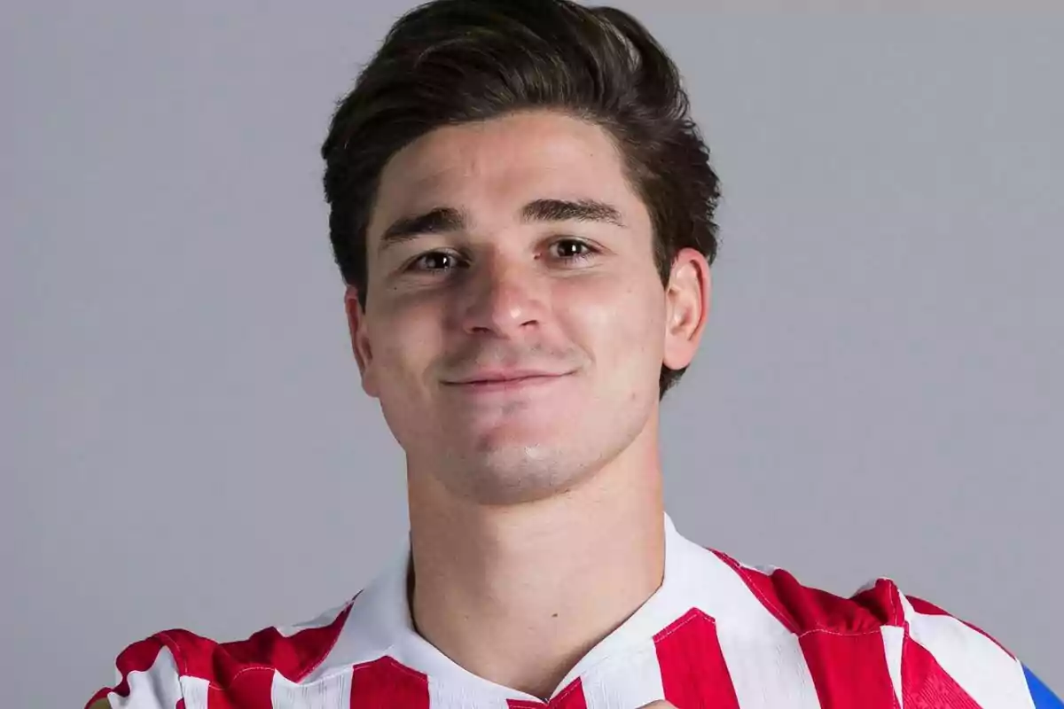 Joven con cabello oscuro y corto viste una camiseta de rayas rojas y blancas y sonríe levemente frente a un fondo gris
