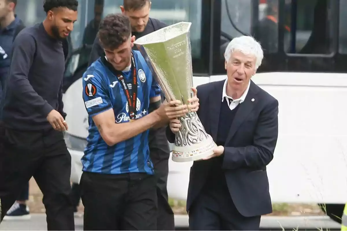 Jugadores y entrenador de un equipo de fútbol celebran sosteniendo un gran trofeo plateado frente a un autobús Jugadores y entrenador de un equipo de fútbol celebran sosteniendo un gran trofeo plateado frente a un autobús