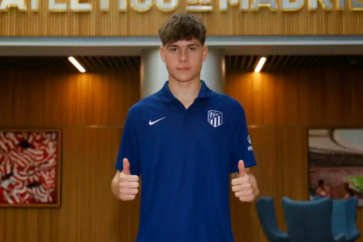 Joven con camiseta azul del Atlético de Madrid posando con ambos pulgares hacia arriba en un interior moderno Joven con camiseta azul del Atlético de Madrid posando con ambos pulgares hacia arriba en un interior moderno