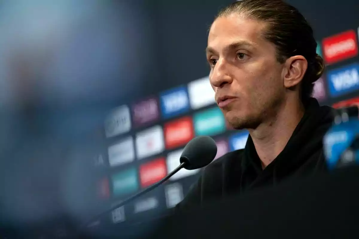 Hombre de cabello recogido hablando frente a un micrófono en una conferencia de prensa con fondo de logotipos desenfocados Hombre de cabello recogido hablando frente a un micrófono en una conferencia de prensa con fondo de logotipos desenfocados
