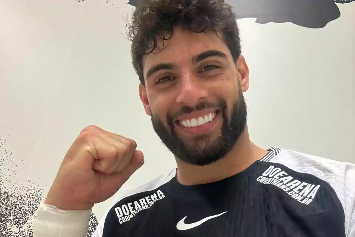 Hombre joven con barba y cabello rizado sonríe y levanta el puño en señal de victoria mientras viste una camiseta deportiva negra y blanca con el logo de Nike y la inscripción Doe Arena Corinthians Hombre joven con barba y cabello rizado sonríe y levanta el puño en señal de victoria mientras viste una camiseta deportiva negra y blanca con el logo de Nike y la inscripción Doe Arena Corinthians