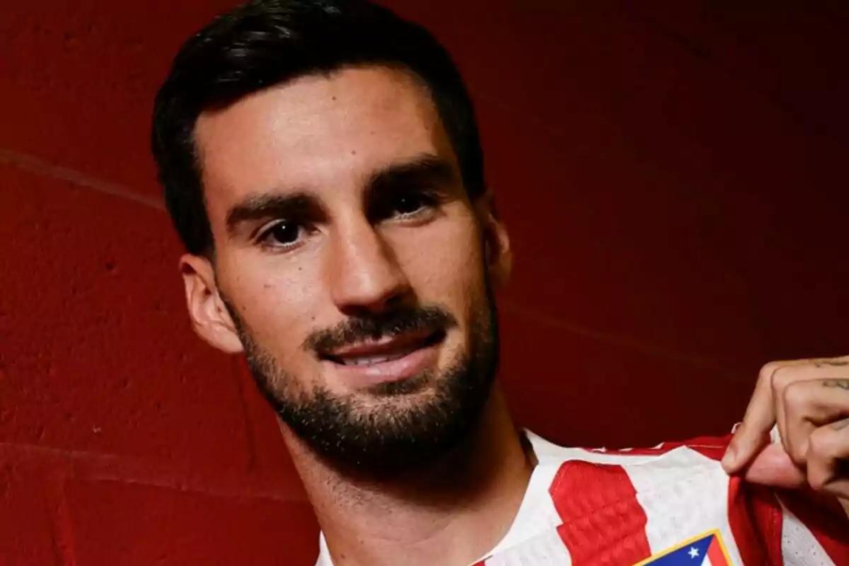 Hombre con barba y cabello oscuro sonríe mientras sostiene la camiseta del Atlético de Madrid frente a una pared roja