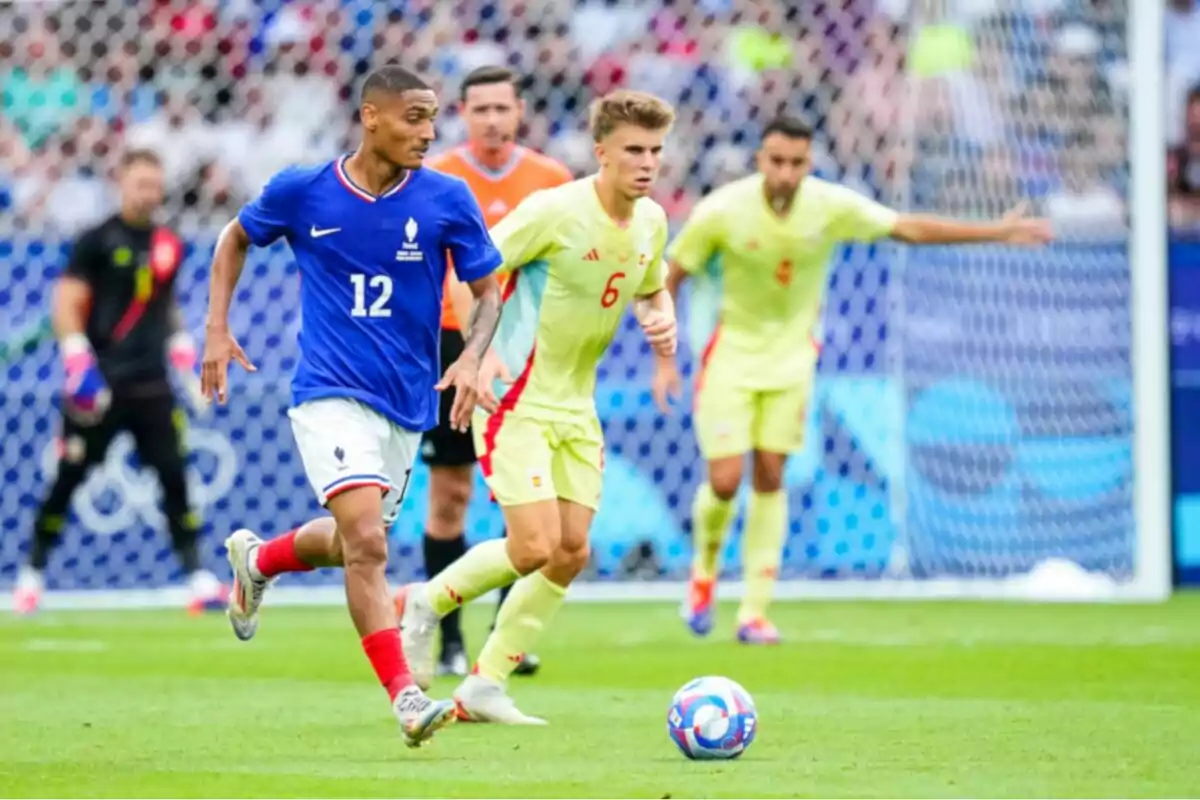 Jugador de fútbol con uniforme azul avanza con el balón mientras es perseguido por un rival con uniforme amarillo en un partido disputado frente a la portería.