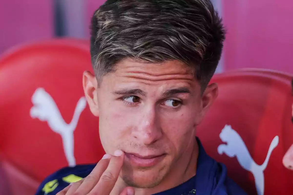 Joven sentado en una banca roja con el logo de Puma al fondo mirando hacia un lado y tocándose la cara