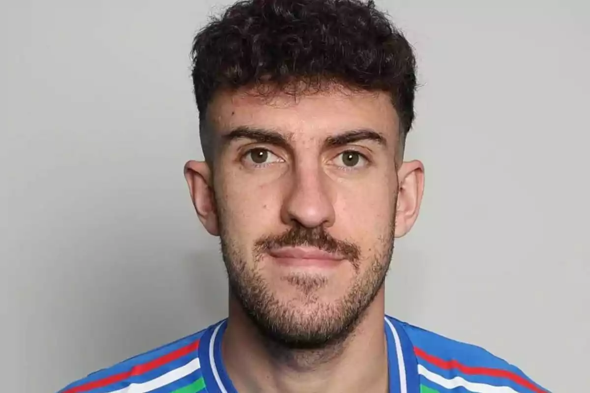 Hombre joven de cabello rizado y barba corta con camiseta deportiva azul mirando al frente sobre fondo gris