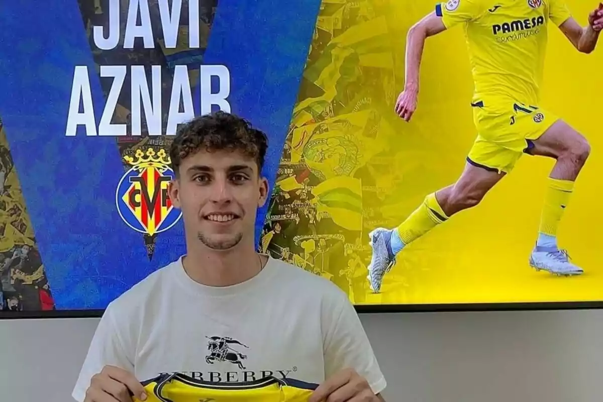 Joven sonriente sostiene una camiseta amarilla frente a una pantalla con su nombre y una imagen de él jugando al fútbol con el uniforme del Villarreal Joven sonriente sostiene una camiseta amarilla frente a una pantalla con su nombre y una imagen de él jugando al fútbol con el uniforme del Villarreal
