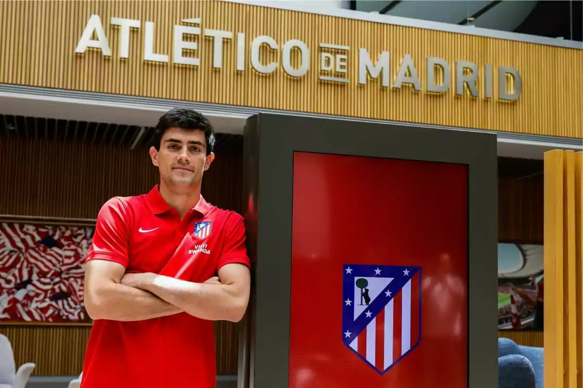 Un hombre con camiseta roja del Atlético de Madrid está de pie con los brazos cruzados frente a un letrero del club y una pantalla con el escudo del equipo Un hombre con camiseta roja del Atlético de Madrid está de pie con los brazos cruzados frente a un letrero del club y una pantalla con el escudo del equipo