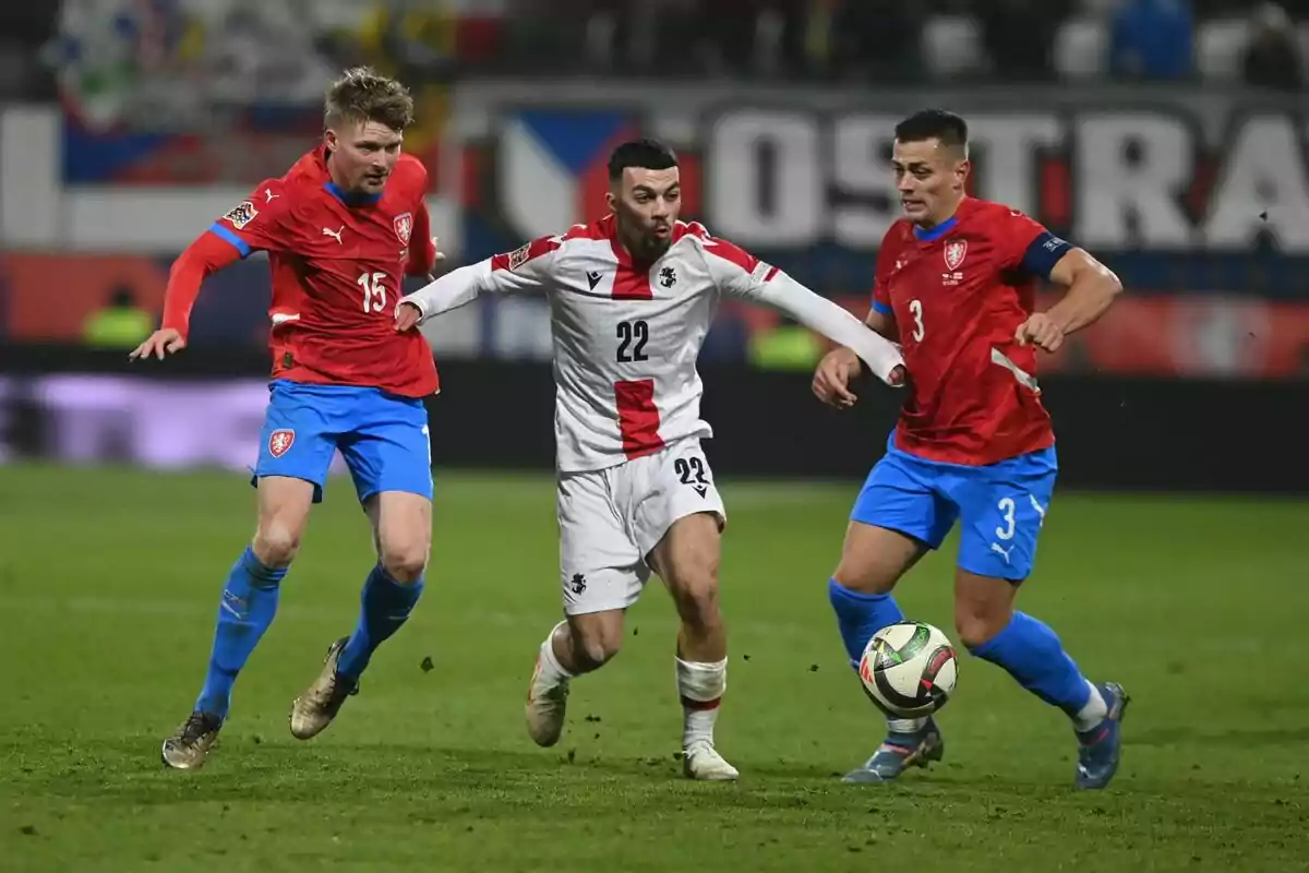 Tres futbolistas disputan el balón durante un partido, dos de ellos con uniforme rojo y azul y uno con uniforme blanco Tres futbolistas disputan el balón durante un partido, dos de ellos con uniforme rojo y azul y uno con uniforme blanco