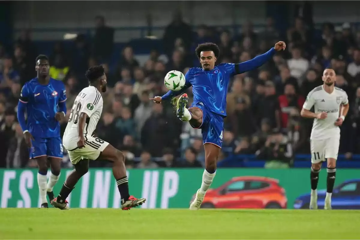 Un jugador de fútbol con uniforme azul controla el balón en el aire mientras un rival con uniforme blanco se acerca durante un partido en un estadio lleno de espectadores. Un jugador de fútbol con uniforme azul controla el balón en el aire mientras un rival con uniforme blanco se acerca durante un partido en un estadio lleno de espectadores.