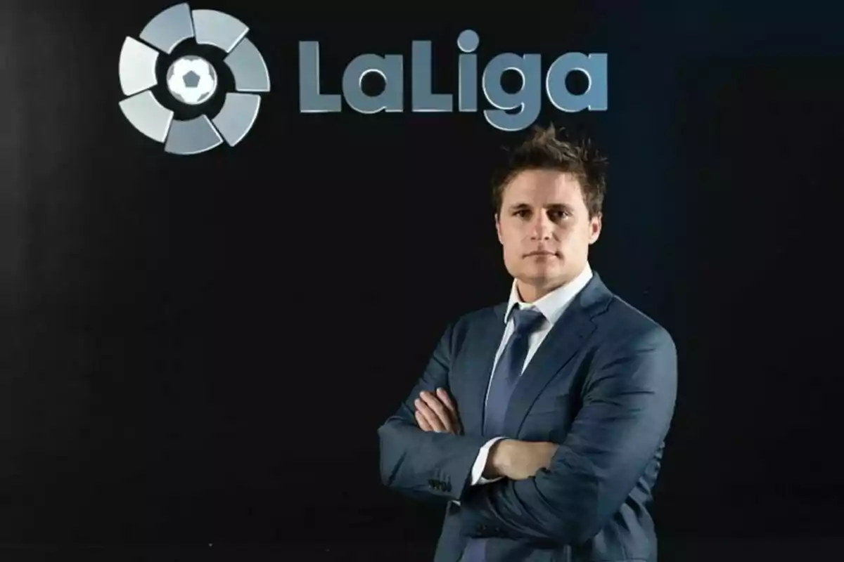 Hombre de traje azul con los brazos cruzados posando frente a un fondo negro con el logo y el texto de LaLiga Hombre de traje azul con los brazos cruzados posando frente a un fondo negro con el logo y el texto de LaLiga