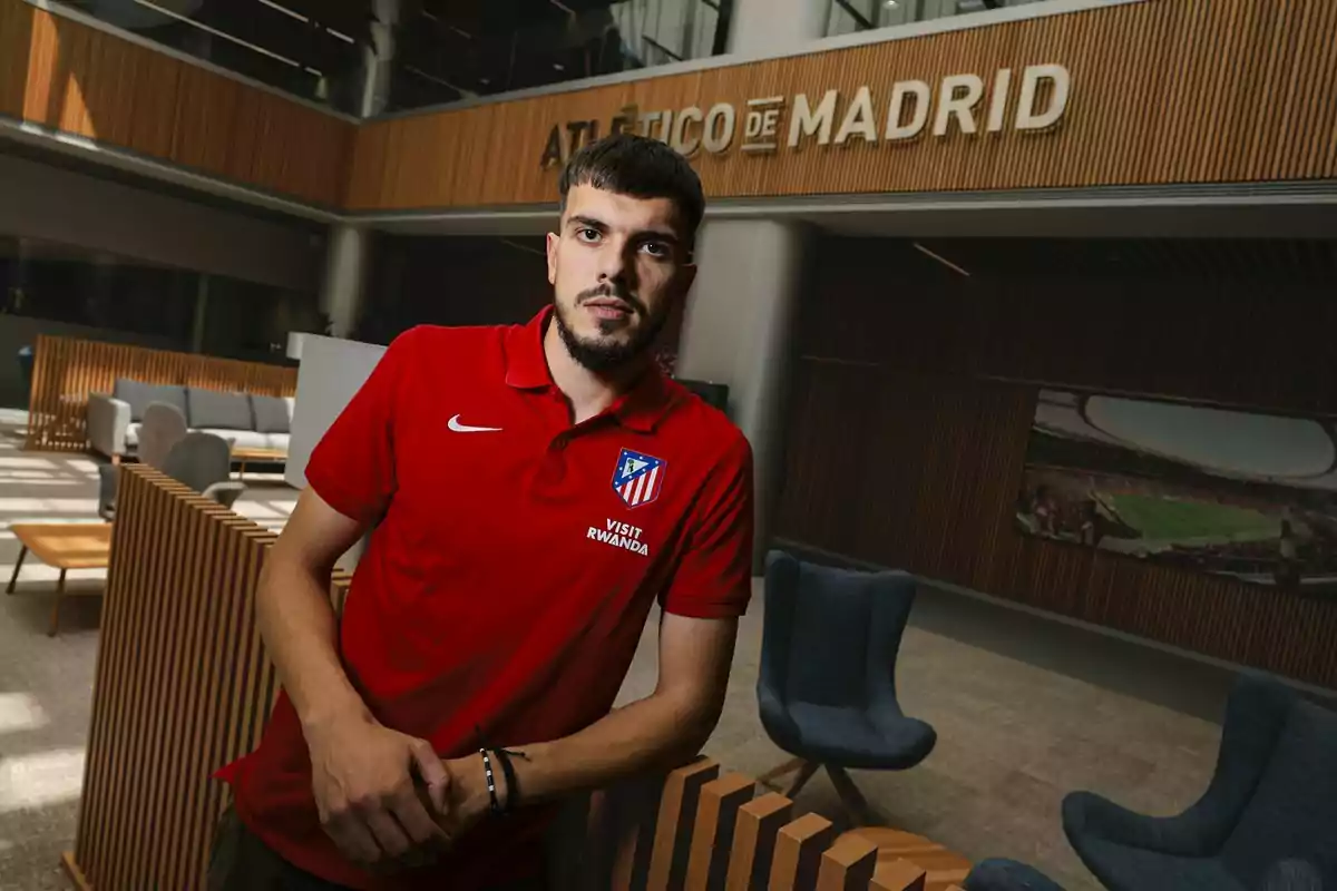 Un hombre con barba y camiseta roja del Atlético de Madrid posa en el interior de un edificio moderno con el letrero Atlético de Madrid al fondo Un hombre con barba y camiseta roja del Atlético de Madrid posa en el interior de un edificio moderno con el letrero Atlético de Madrid al fondo