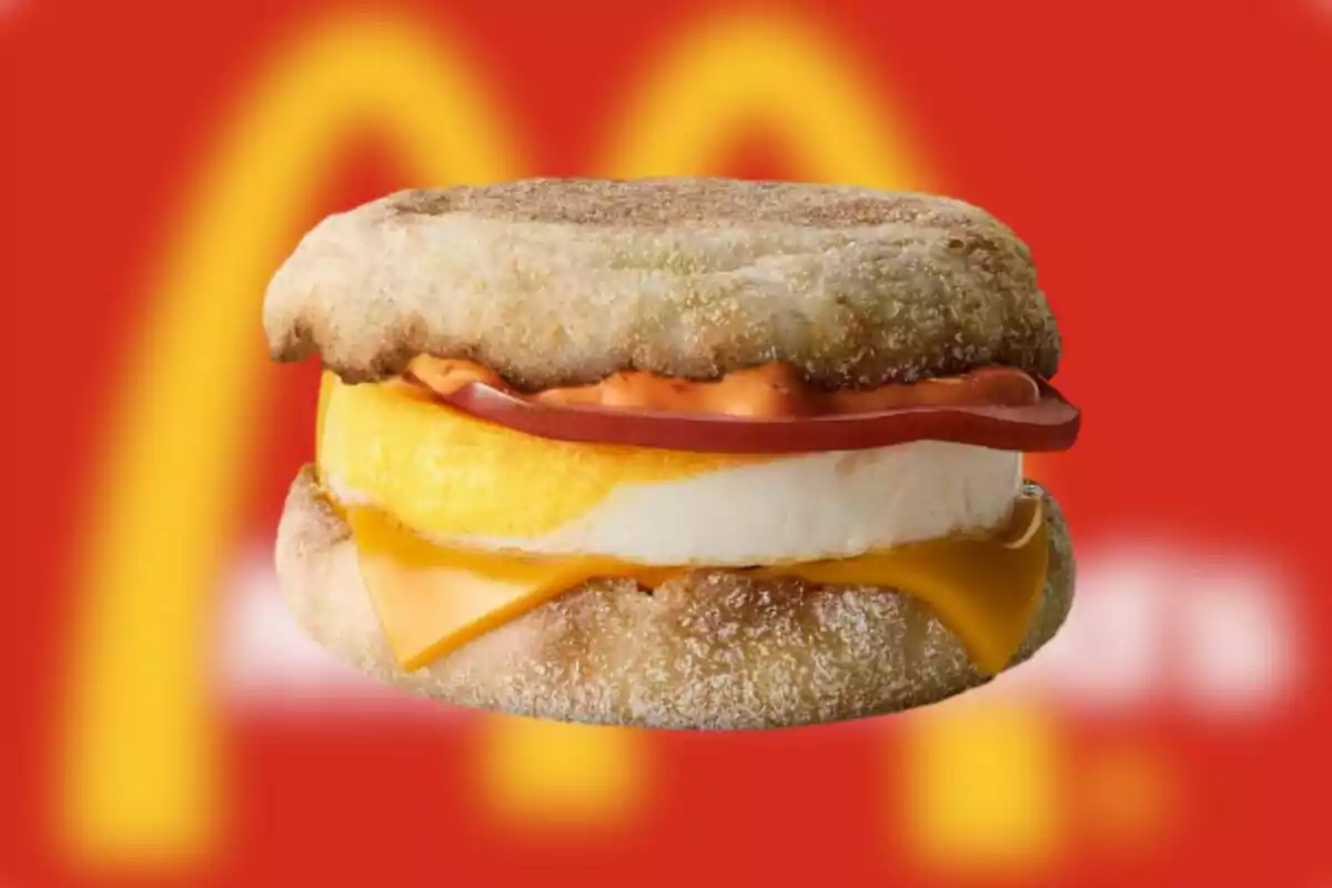 Sándwich de desayuno con huevo, queso, jamón y pan inglés sobre un fondo rojo con el logo de McDonald's desenfocado Sándwich de desayuno con huevo, queso, jamón y pan inglés sobre un fondo rojo con el logo de McDonald's desenfocado
