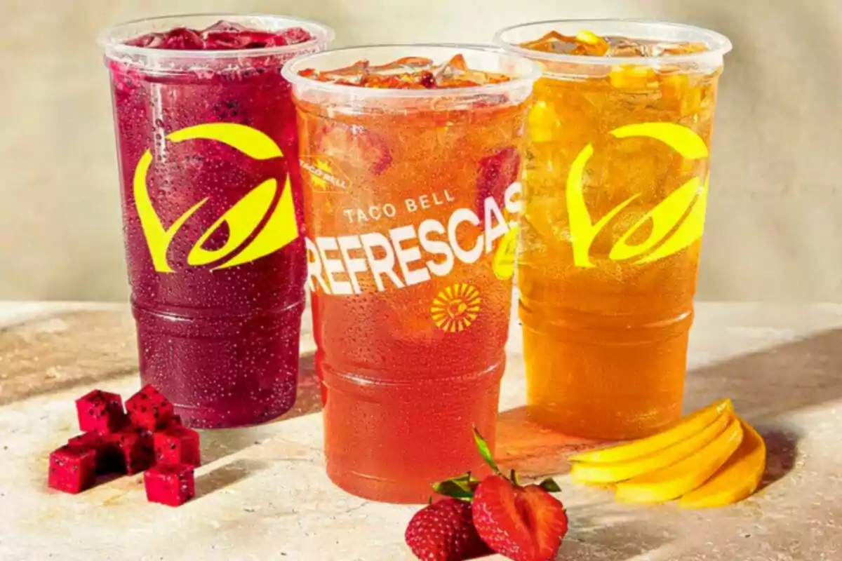 Three large glasses of colorful soft drinks—red, orange, and yellow—with ice, accompanied by pieces of fruit such as strawberry, mango, and dragon fruit, on a light surface. Three large glasses of colorful soft drinks—red, orange, and yellow—with ice, accompanied by pieces of fruit such as strawberry, mango, and dragon fruit, on a light surface.