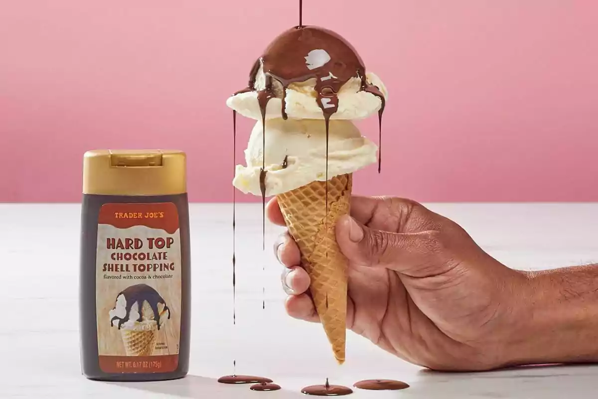 Una mano sostiene un cono de helado de vainilla sobre el que se vierte cobertura de chocolate líquido, junto a un envase de topping de chocolate sobre una mesa blanca con fondo rosa. Una mano sostiene un cono de helado de vainilla sobre el que se vierte cobertura de chocolate líquido, junto a un envase de topping de chocolate sobre una mesa blanca con fondo rosa.