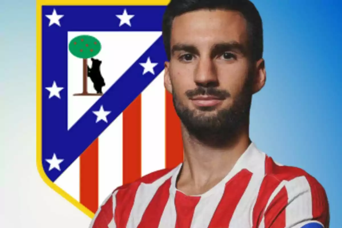Jugador con camiseta rojiblanca posando frente al escudo del Atlético de Madrid sobre fondo azul.
