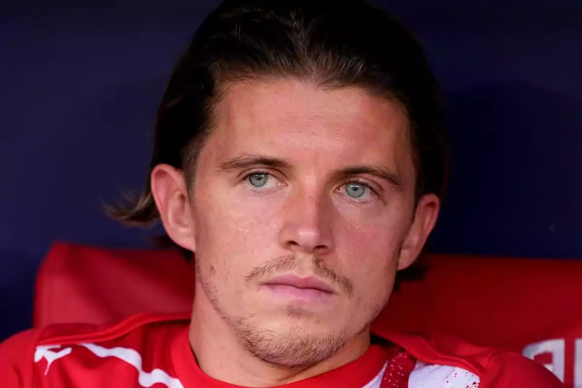 Hombre joven con cabello castaño y ojos claros sentado con expresión seria y camiseta roja