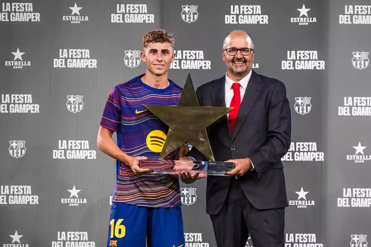Un futbolista del Barcelona y un hombre con traje sostienen juntos un trofeo en forma de estrella frente a un fondo con el logo de La Festa del Gamper. Un futbolista del Barcelona y un hombre con traje sostienen juntos un trofeo en forma de estrella frente a un fondo con el logo de La Festa del Gamper.