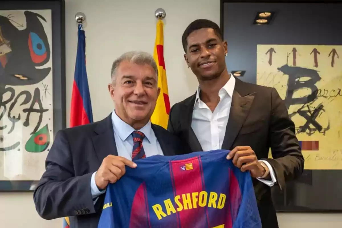 Dos hombres posan sonrientes mientras sostienen una camiseta del FC Barcelona con el nombre Rashford en un despacho decorado con banderas y cuadros del club. Dos hombres posan sonrientes mientras sostienen una camiseta del FC Barcelona con el nombre Rashford en un despacho decorado con banderas y cuadros del club.
