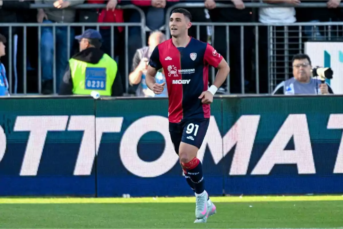Futbolista con uniforme azul y rojo corriendo en el campo durante un partido con público y fotógrafos al fondo Futbolista con uniforme azul y rojo corriendo en el campo durante un partido con público y fotógrafos al fondo