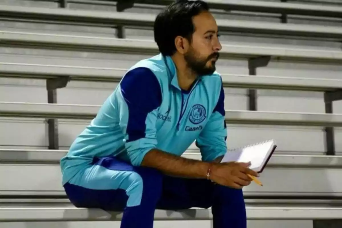 Hombre con uniforme deportivo azul sentado en gradas sosteniendo una libreta y un lápiz mientras observa atentamente. Hombre con uniforme deportivo azul sentado en gradas sosteniendo una libreta y un lápiz mientras observa atentamente.