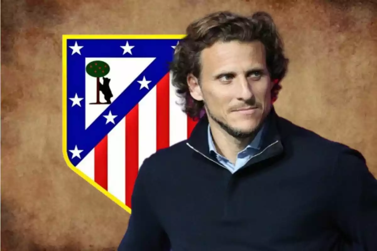 Hombre de cabello rizado y suéter oscuro posando frente al escudo del Atlético de Madrid sobre un fondo marrón.