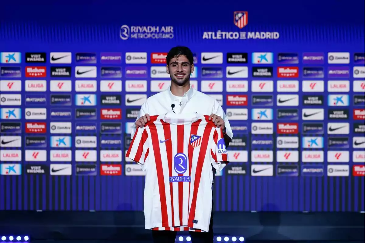 Un jugador sonriente sostiene la camiseta del Atlético de Madrid durante su presentación oficial frente a un fondo con logotipos de patrocinadores y el escudo del club. Un jugador sonriente sostiene la camiseta del Atlético de Madrid durante su presentación oficial frente a un fondo con logotipos de patrocinadores y el escudo del club.