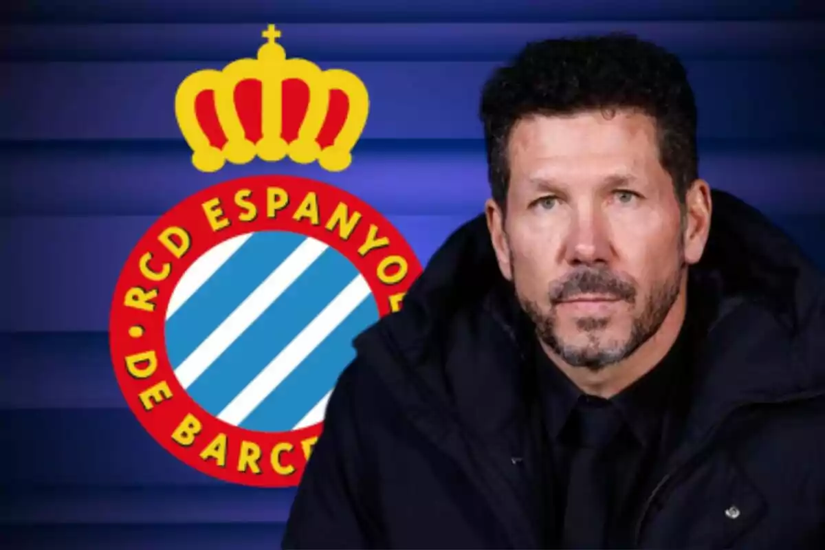 Hombre con barba y cabello corto oscuro vistiendo abrigo negro frente al escudo del RCD Espanyol de Barcelona sobre un fondo azul