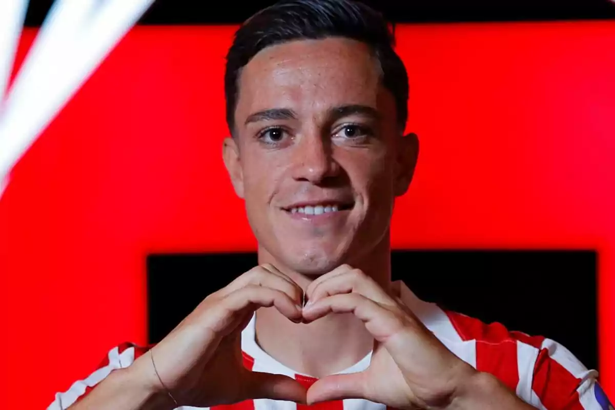 Jugador de fútbol con camiseta de rayas rojas y blancas haciendo un gesto de corazón con las manos frente a un fondo rojo