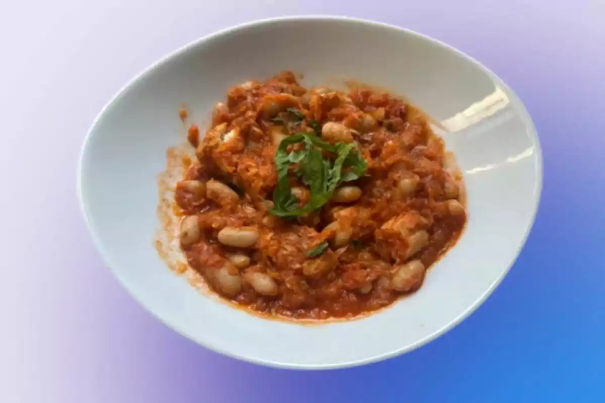 Plato hondo con guiso de alubias blancas en salsa de tomate decorado con hojas verdes sobre fondo degradado azul y lila Plato hondo con guiso de alubias blancas en salsa de tomate decorado con hojas verdes sobre fondo degradado azul y lila