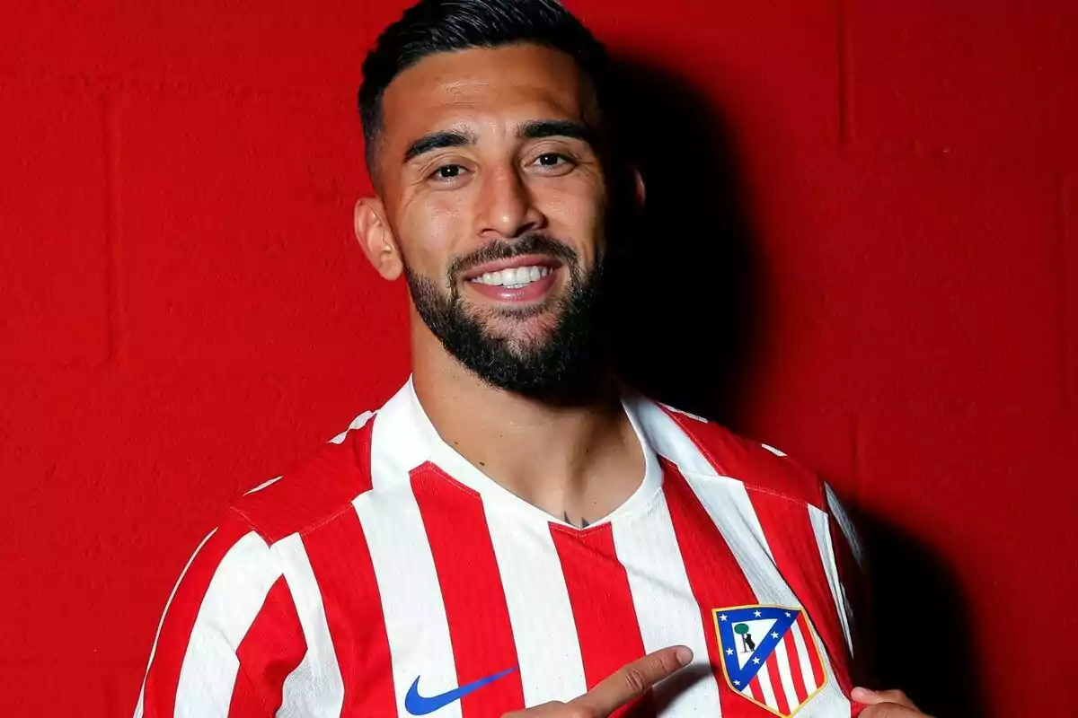 Futbolista sonriente con barba y cabello corto señalando el escudo del Atlético de Madrid en su camiseta frente a una pared roja Futbolista sonriente con barba y cabello corto señalando el escudo del Atlético de Madrid en su camiseta frente a una pared roja