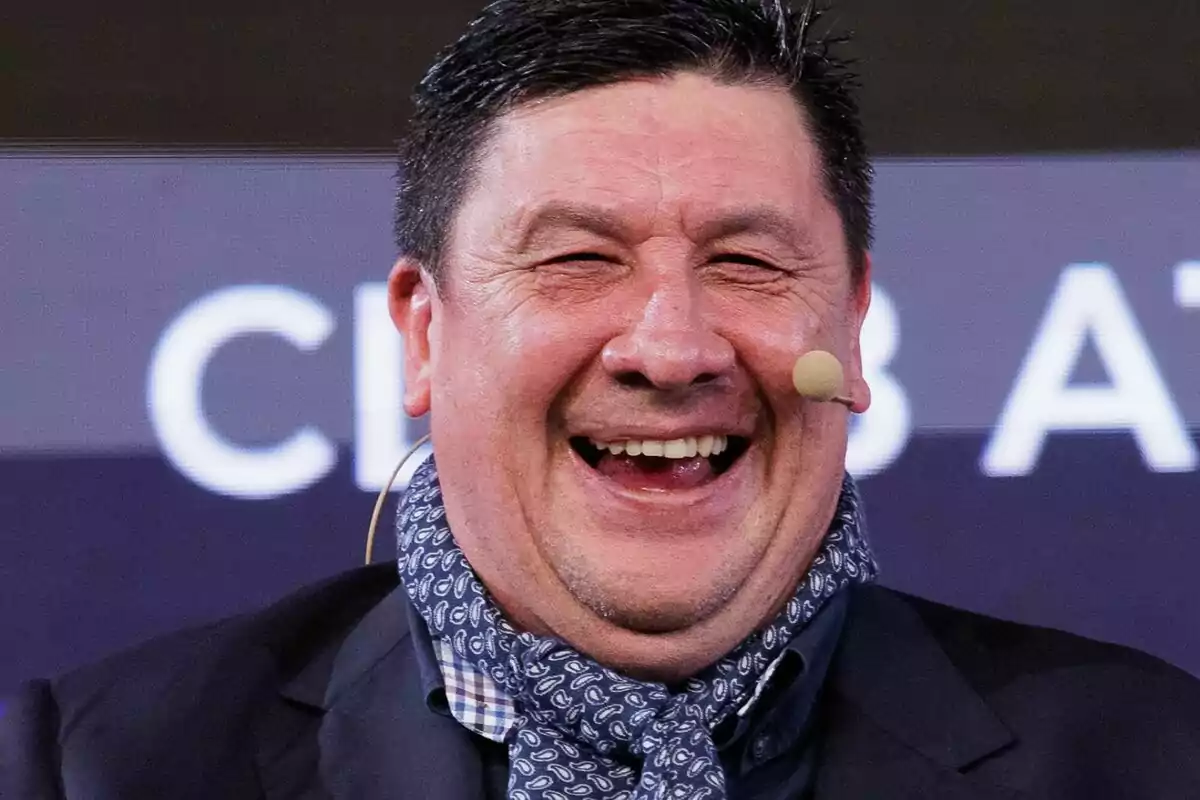 Hombre de mediana edad sonriendo ampliamente con micrófono en la mejilla y bufanda azul