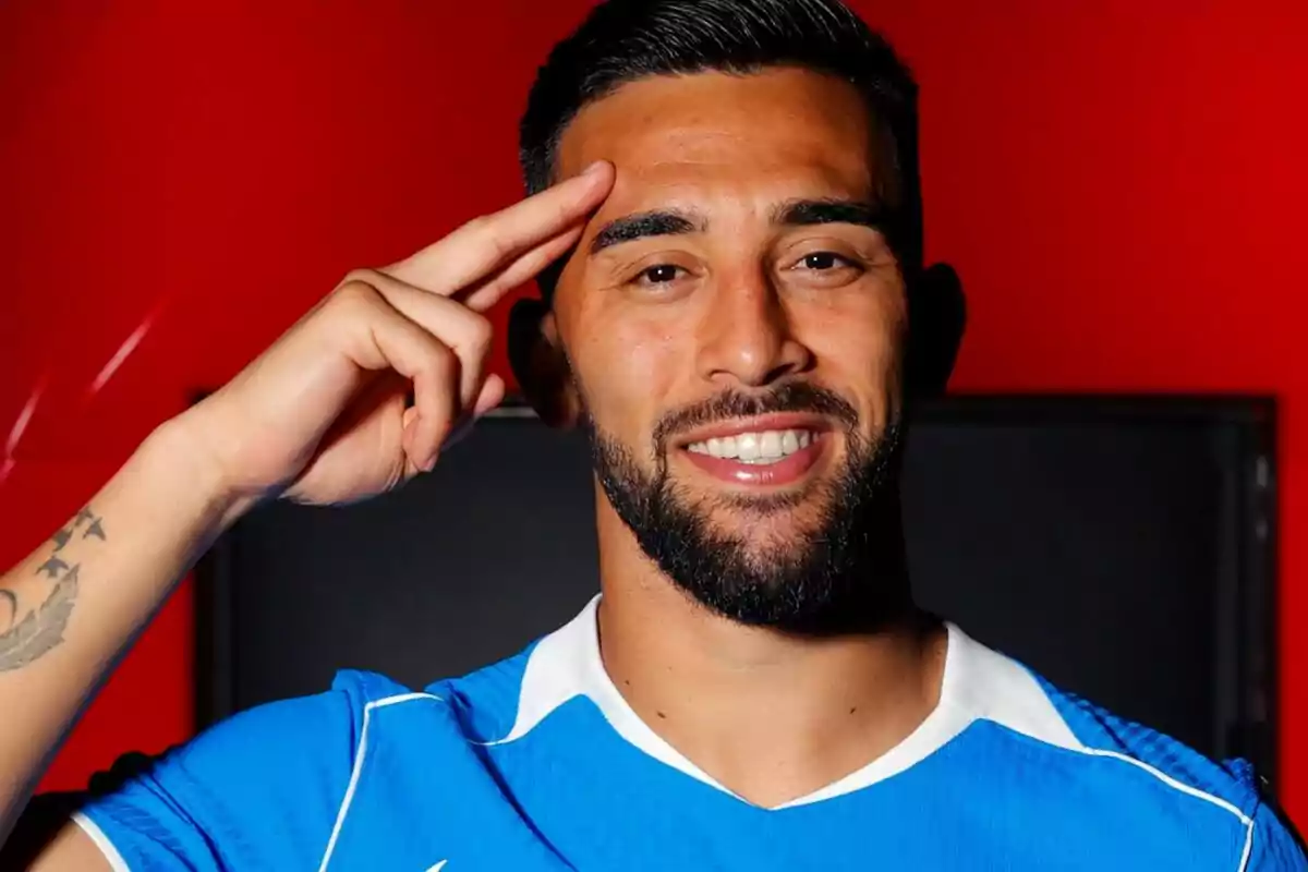 Hombre con barba y camiseta azul sonriendo y haciendo un saludo militar frente a un fondo rojo