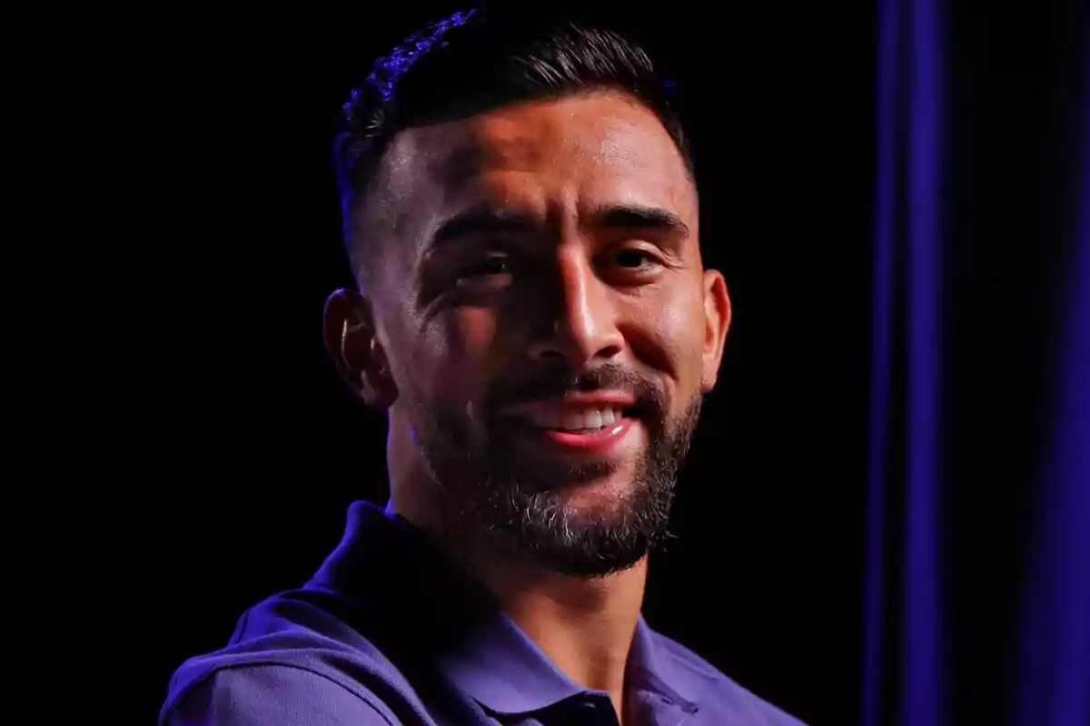 Hombre joven con barba y cabello corto sonriendo, vestido con camisa polo morada y fondo oscuro