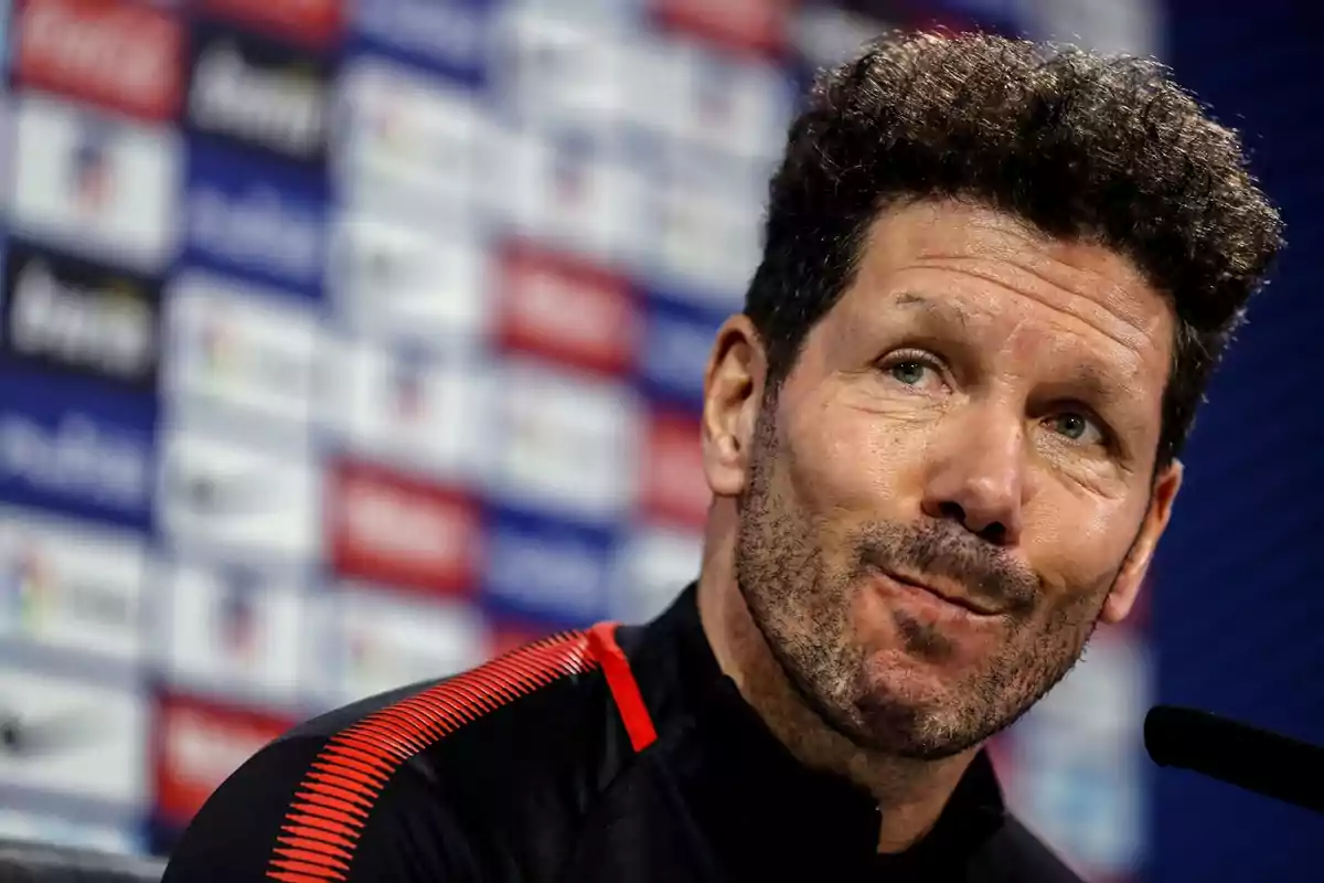 Un hombre con barba y cabello rizado en una conferencia de prensa con un fondo de logotipos.