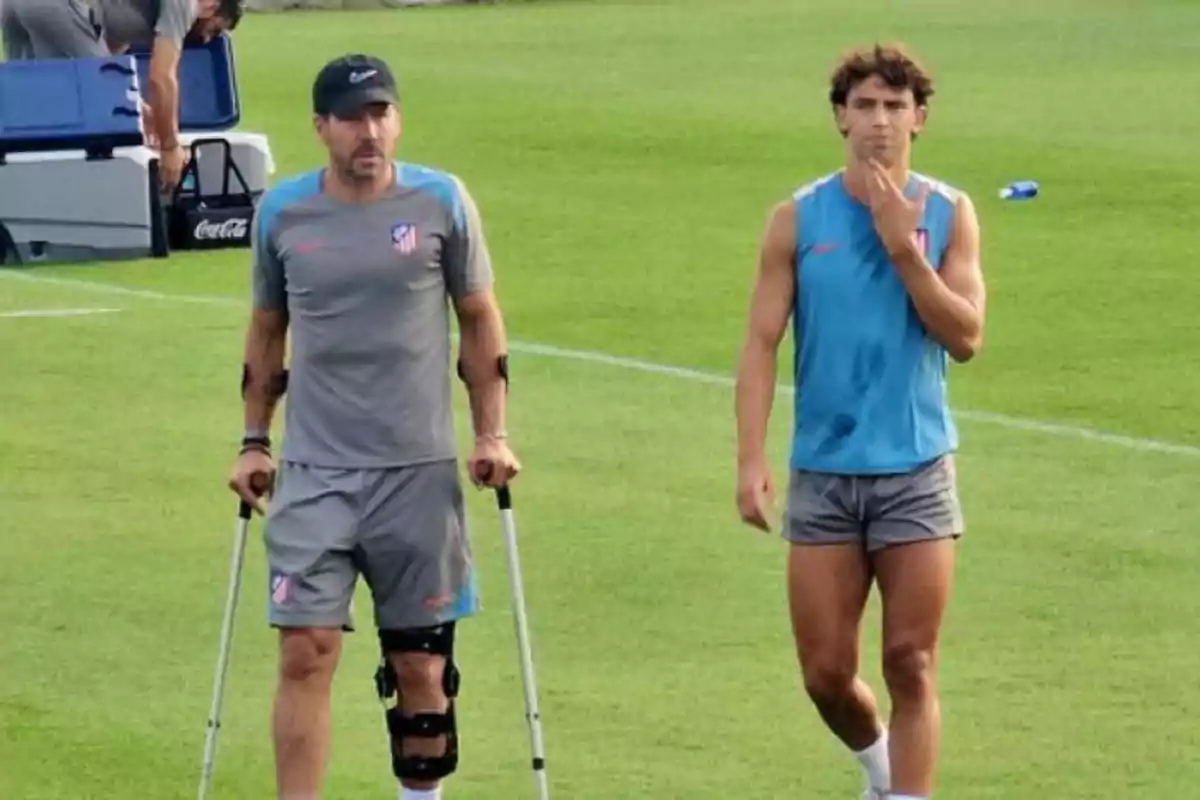 Dos personas en un campo de fútbol, una de ellas con muletas y una rodillera, ambas con ropa deportiva del Atlético de Madrid.