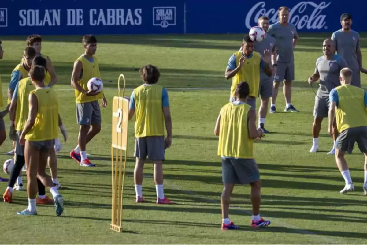 Jugadores de fútbol entrenando en un campo de fútbol con petos amarillos y entrenadores observando.