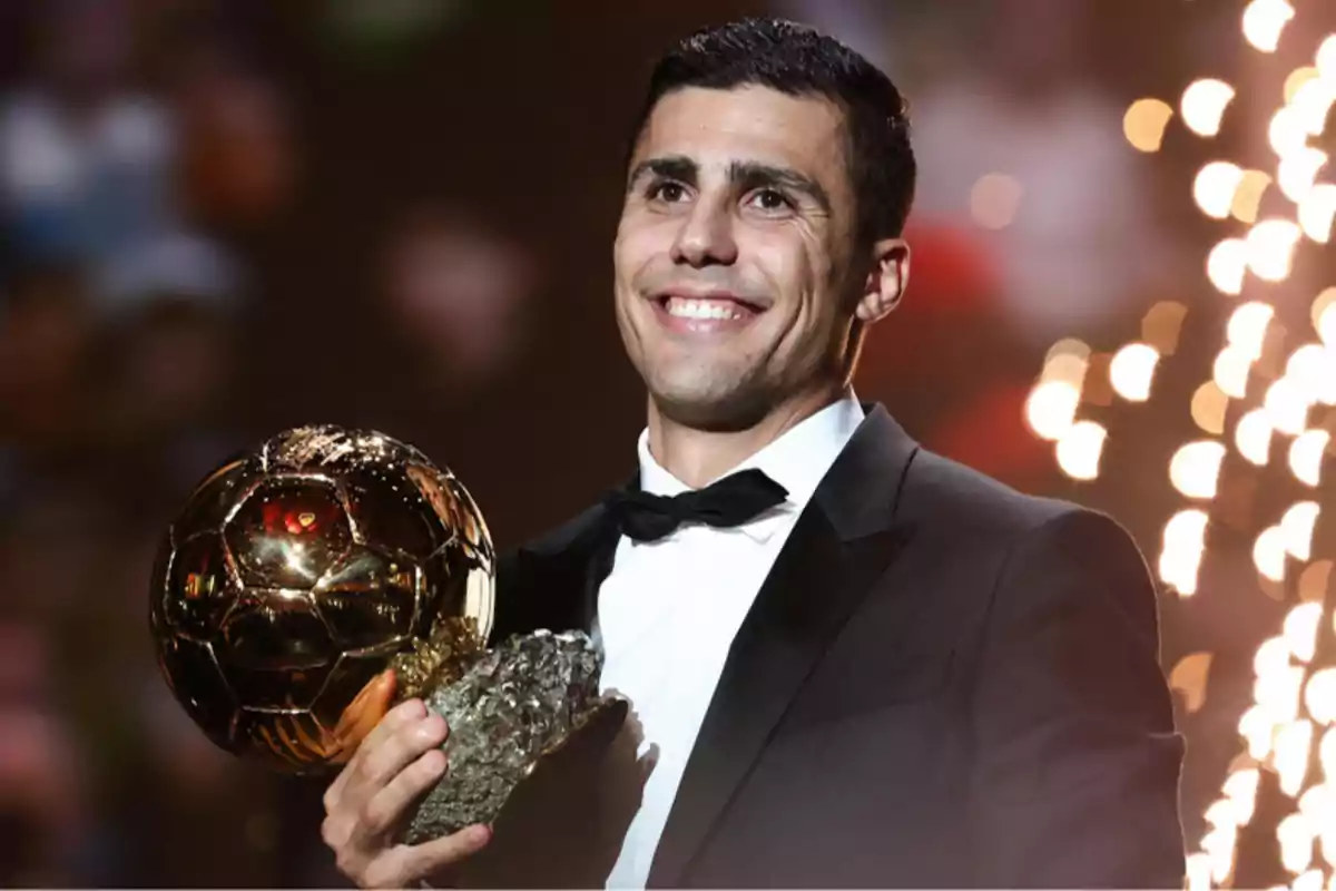 Un hombre sonriente en traje sostiene un trofeo dorado con luces brillantes de fondo.