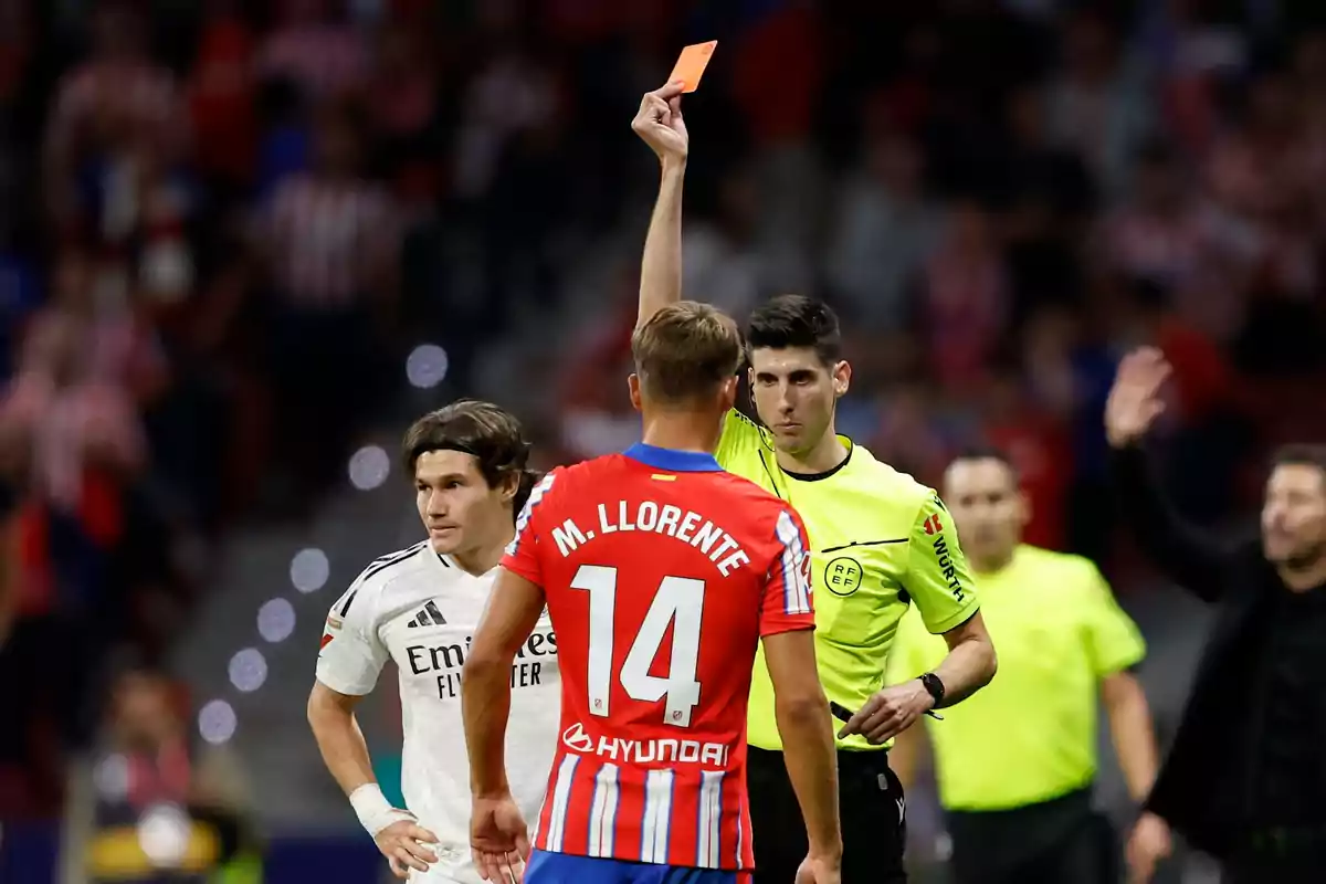 Un árbitro muestra una tarjeta roja a un jugador de fútbol con el número 14 en su camiseta mientras otro jugador observa la escena.