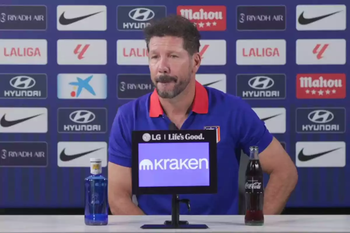 Un hombre con barba y bigote, vestido con una camiseta azul con cuello rojo, está sentado en una mesa durante una conferencia de prensa. Frente a él hay una botella de agua y una botella de Coca-Cola. En el fondo se ven varios logotipos de patrocinadores como Hyundai, Nike, Mahou y LaLiga.