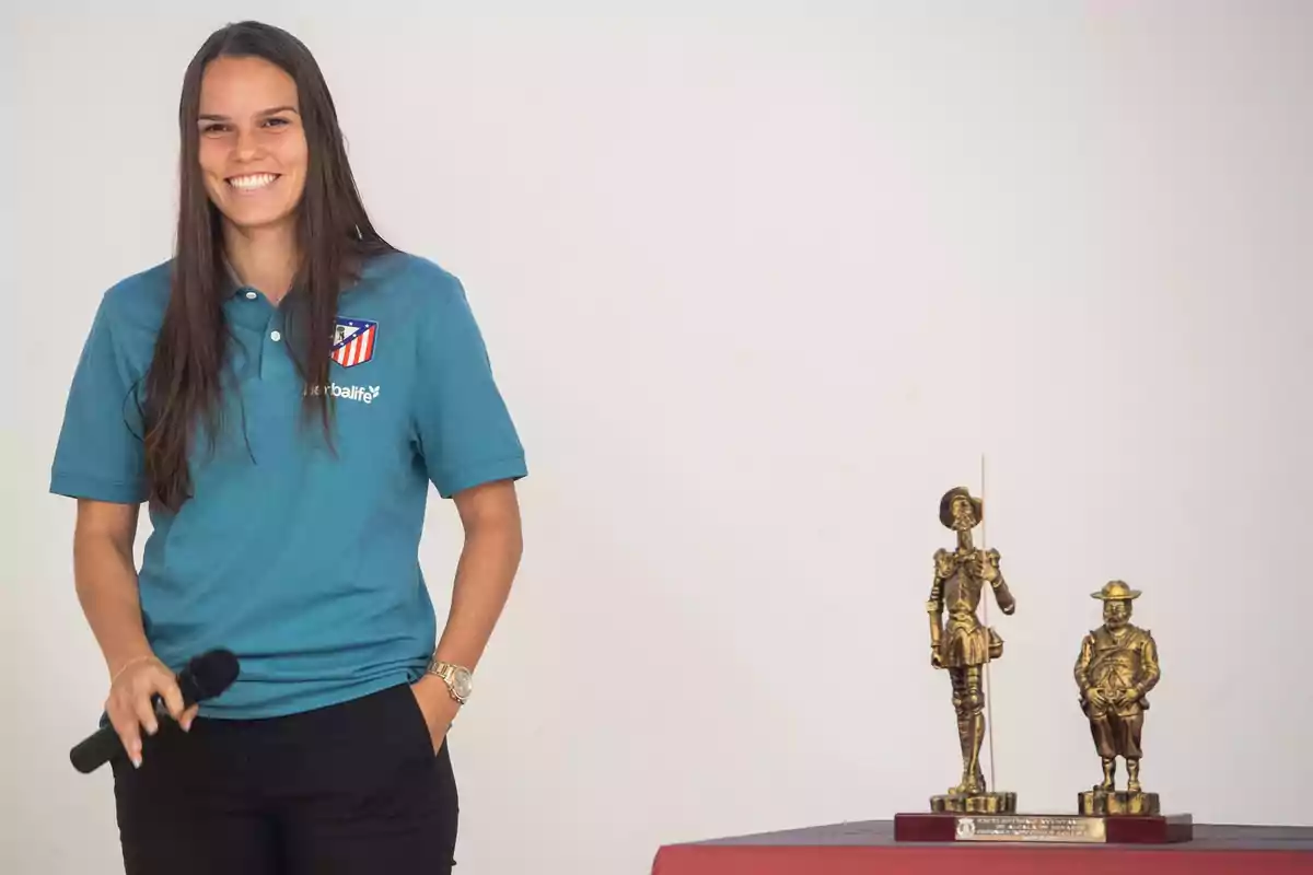 Una mujer sonriente con una camiseta del Atlético de Madrid sostiene un micrófono mientras está de pie junto a dos estatuillas doradas sobre una mesa.